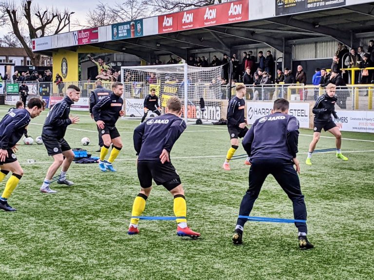 Players performing warmup exercises on a football field in England with spectators watching.